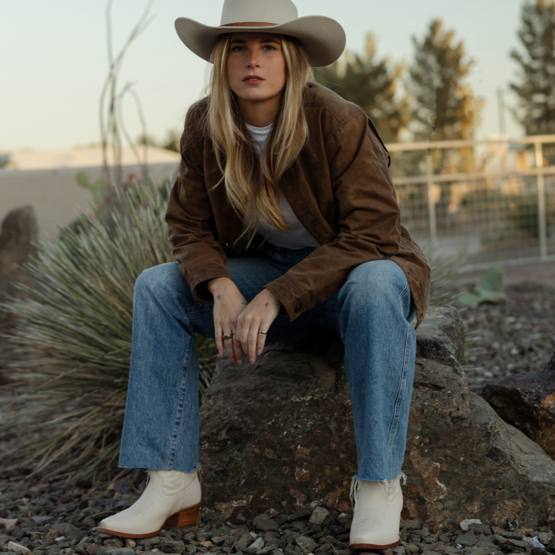 Women sitting on rock with white cowboy boots, blue jeans, brown coat and off white cowboy hat