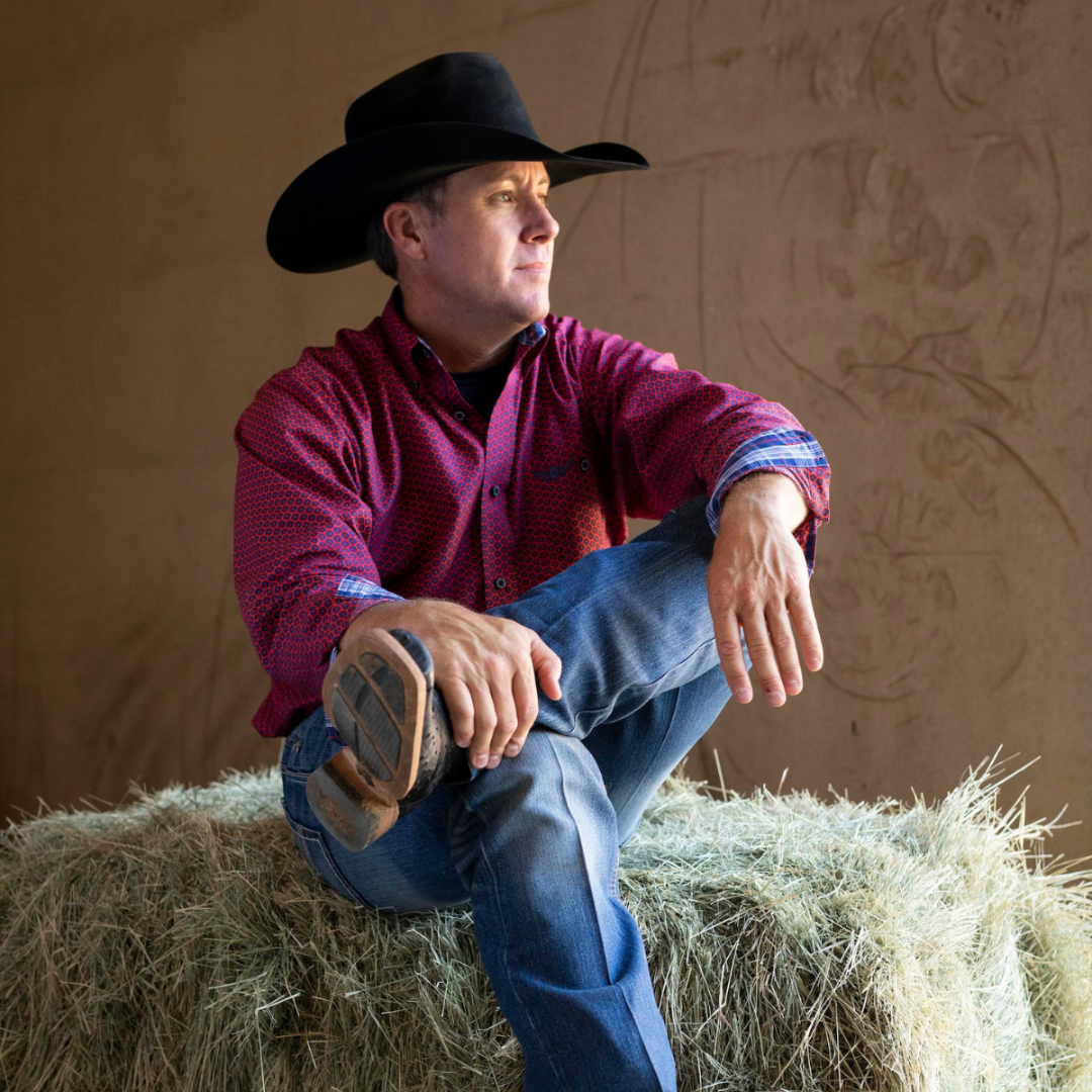 Man sitting on hay with blue jeans, black cowboy hat and brown boots