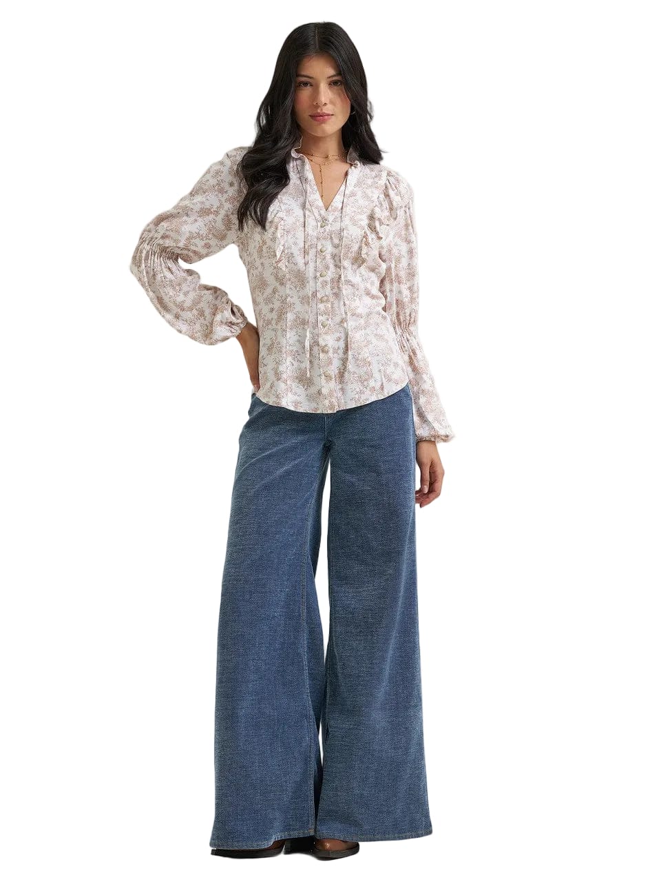 A woman modeling a long-sleeve button-up blouse with a light beige and white floral pattern, paired with wide-leg blue jeans, against a plain light gray studio background.