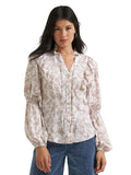 A woman modeling a long-sleeve blouse with a beige and white floral pattern, ruffled collar and cuffs, and a button-down front, paired with blue jeans against a light gray studio background.