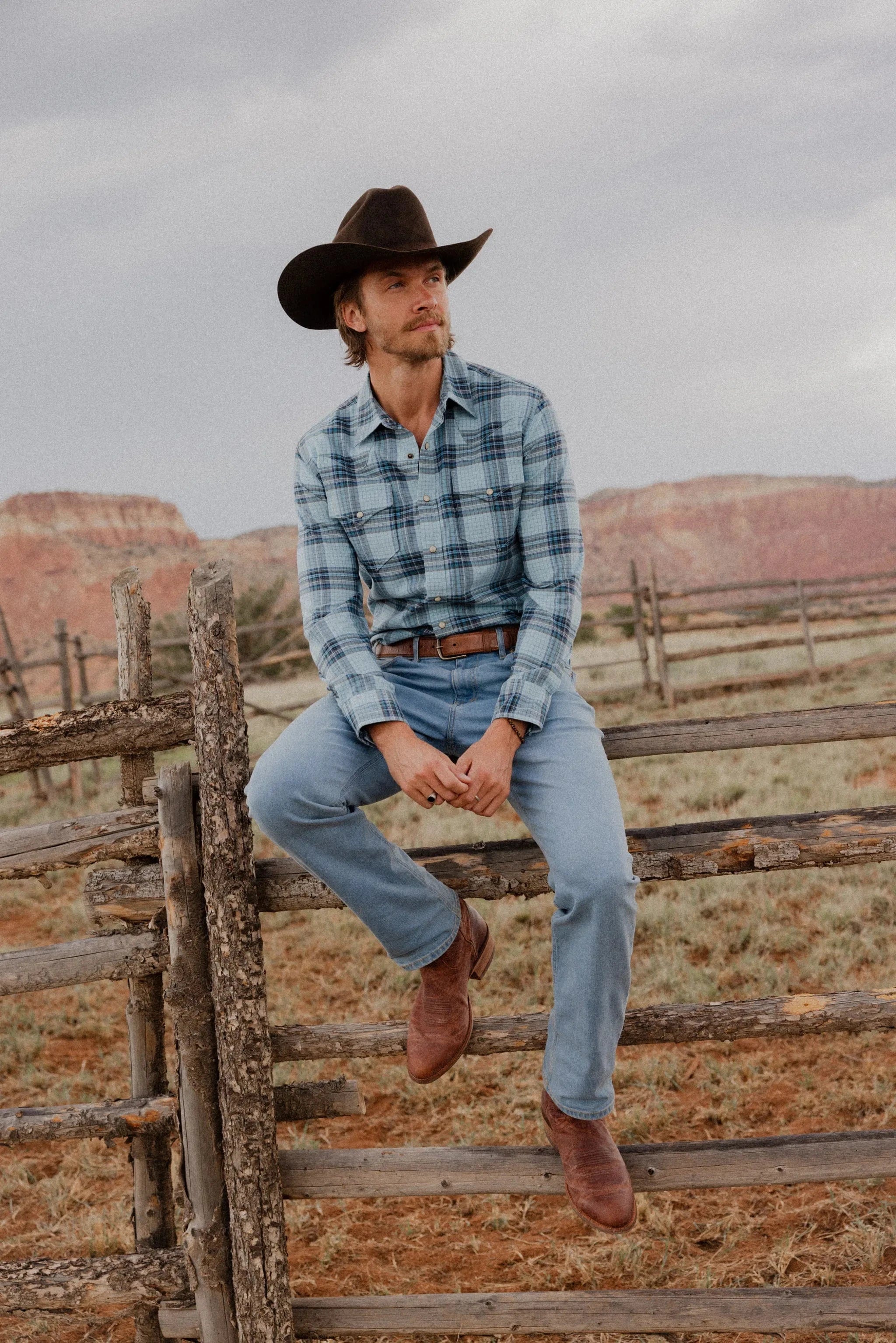 Man in cowboy hat and plaid shirt sitting on a wooden fence with desert landscape in the background