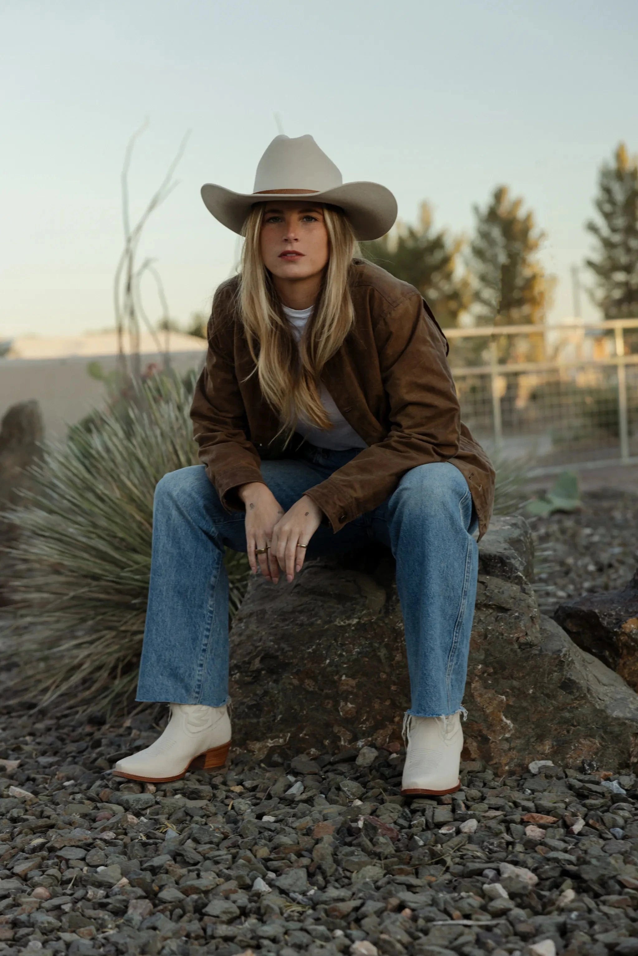 Person wearing a cowboy hat, brown jacket, blue jeans, and white boots sitting on a rock.